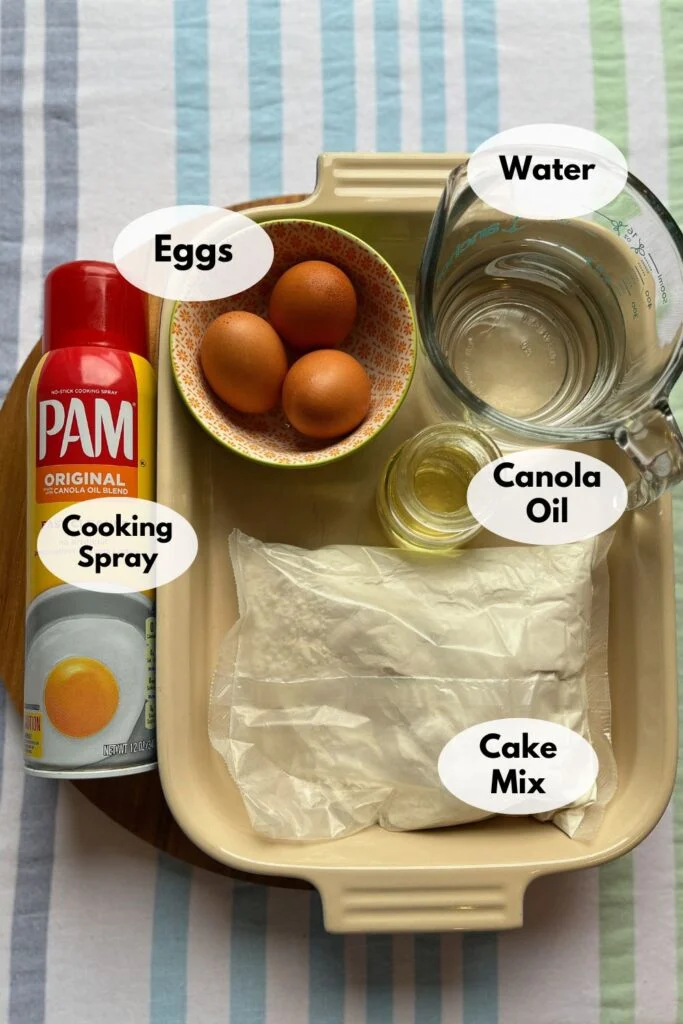 A baking dish containing three eggs, a measuring cup with water, a small cup of canola oil, a bottle of cooking spray, and a bag of cake mix, all on a striped tablecloth.