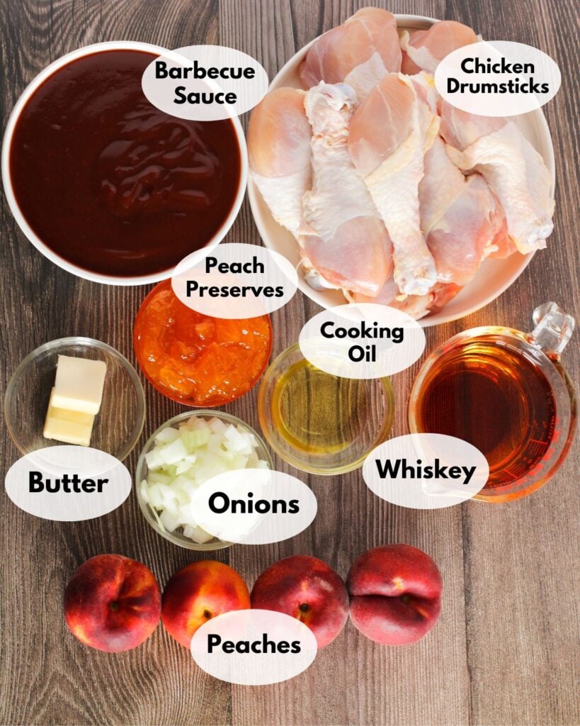 Ingredients for a recipe including chicken drumsticks, barbecue sauce, peach preserves, cooking oil, whiskey, butter, onions, and peaches are displayed on a wooden surface.