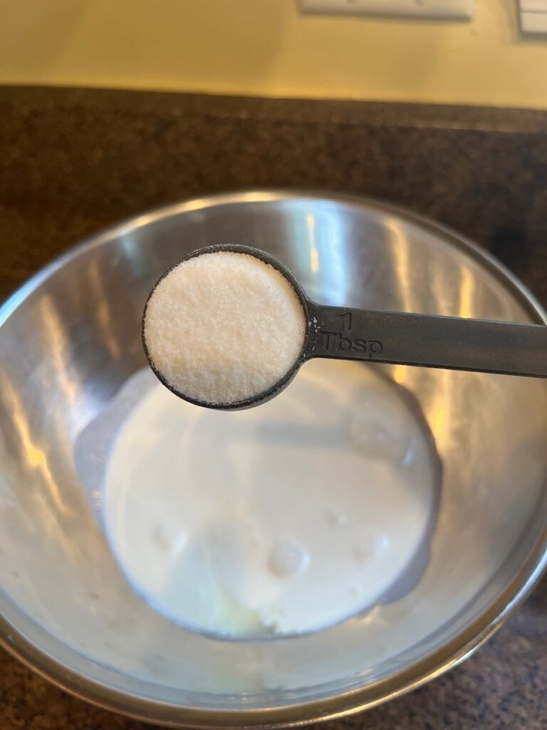 A tablespoon of white powder is held above a stainless steel mixing bowl containing a liquid mixture.