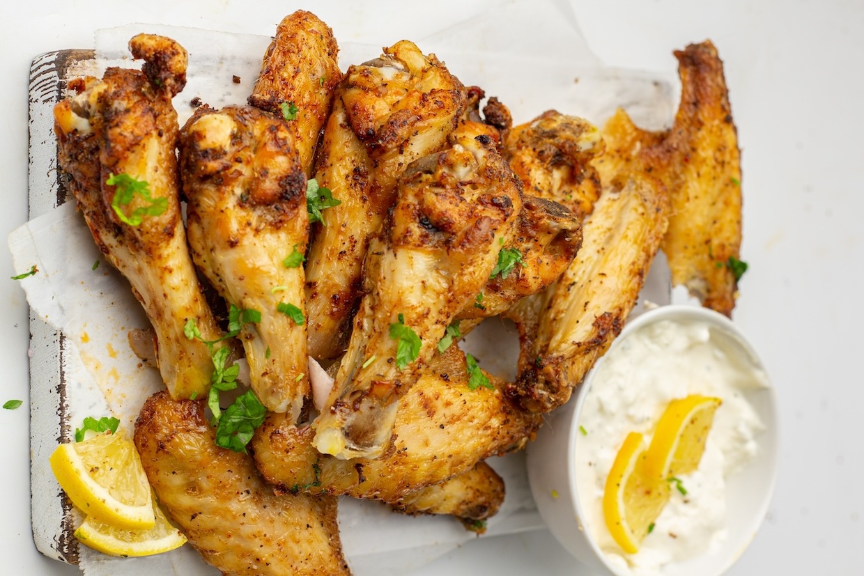 A pile of seasoned, grilled chicken wings on a wooden board, garnished with chopped herbs, alongside a bowl of white dipping sauce with lemon wedges.