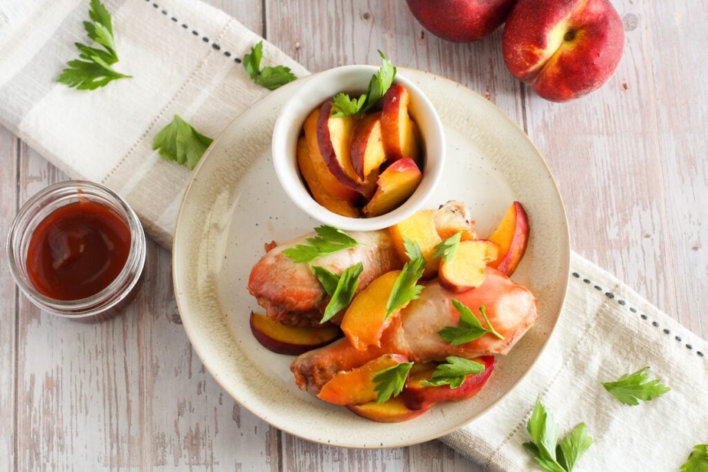 A plate of cooked chicken drumsticks garnished with fresh herbs and sliced peaches. A small bowl of sliced peaches and a small jar of red sauce are on the side, with a cloth and two whole peaches nearby.