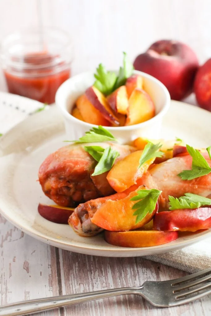 A plate with cooked chicken drumsticks garnished with fresh parsley and sliced peaches, accompanied by a small bowl of additional peach slices. A jar of sauce and whole peaches are in the background.