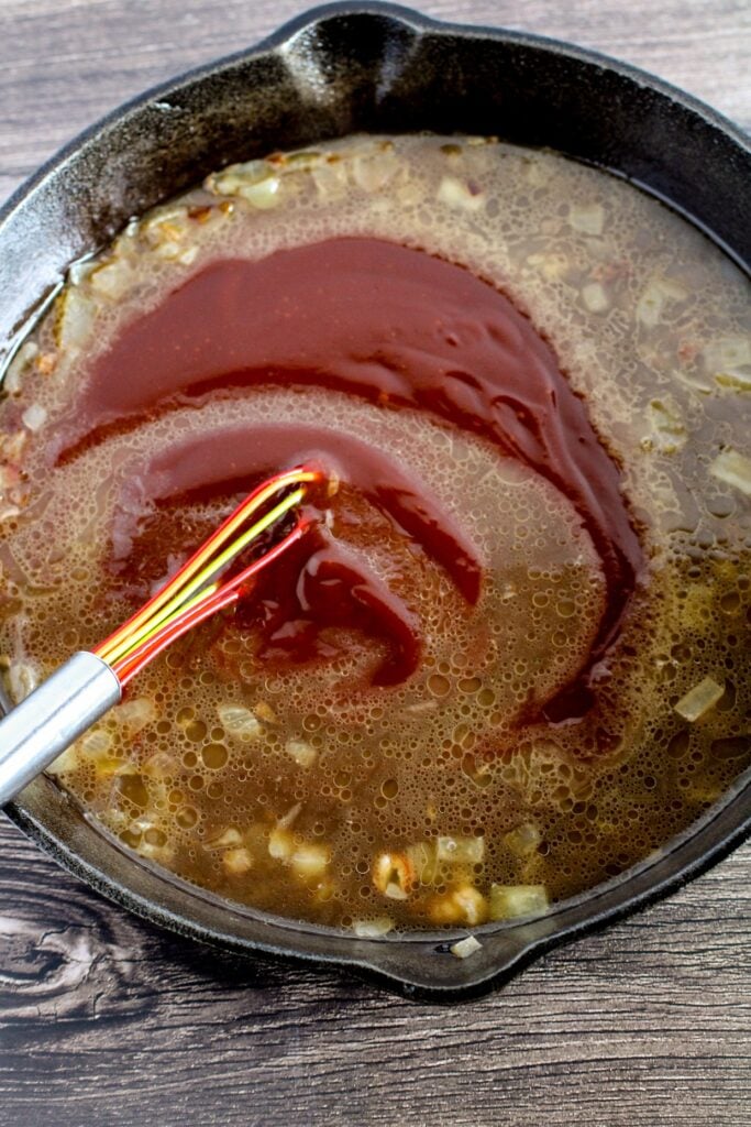 A metal whisk stirring red sauce into a pan of broth containing onions and other small ingredients.