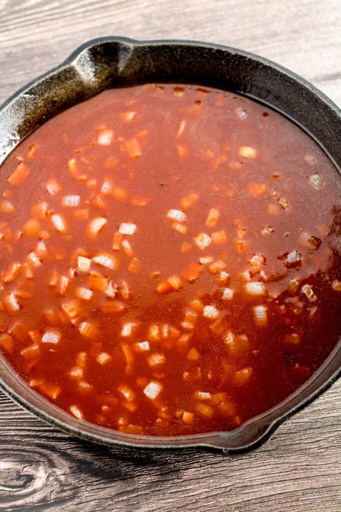 A cast iron skillet filled with a tomato-based sauce containing chopped onions, resting on a wooden surface.