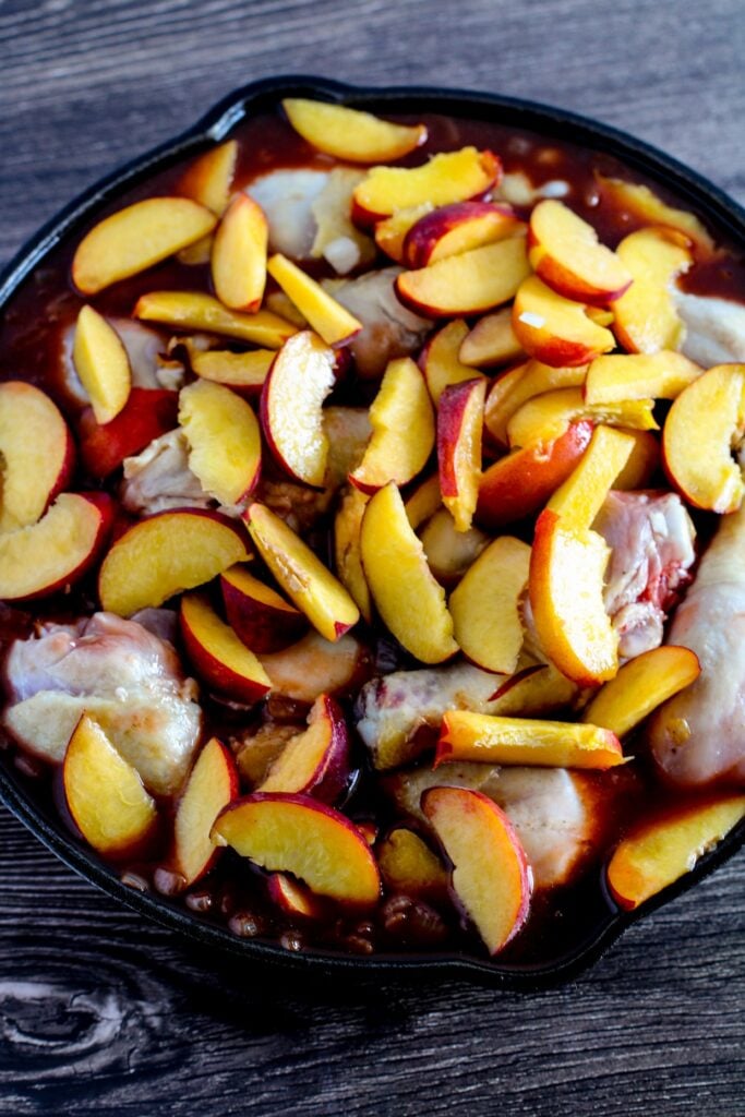 A skillet containing raw chicken pieces covered with sliced peaches and a thick sauce, placed on a dark wooden surface.