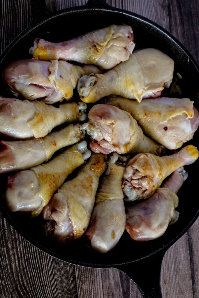A cast iron skillet filled with raw chicken drumsticks, arranged in a circular pattern.