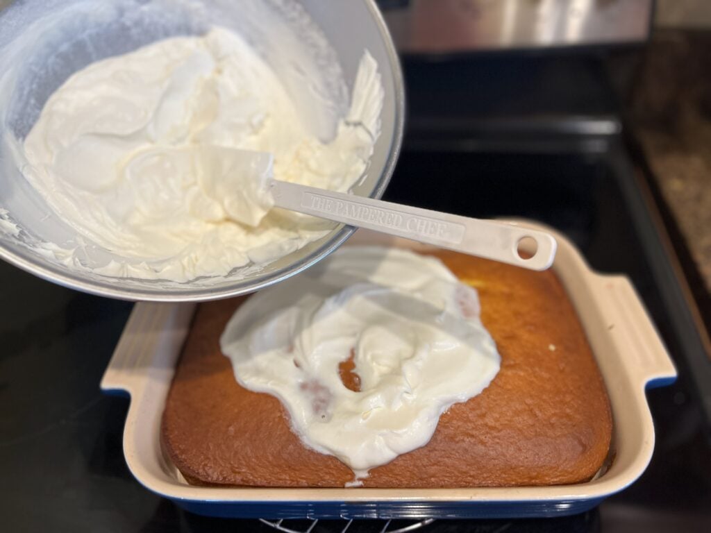A spatula spreads a thick layer of white frosting from a metal bowl onto a square-shaped baked cake placed in a ceramic dish.