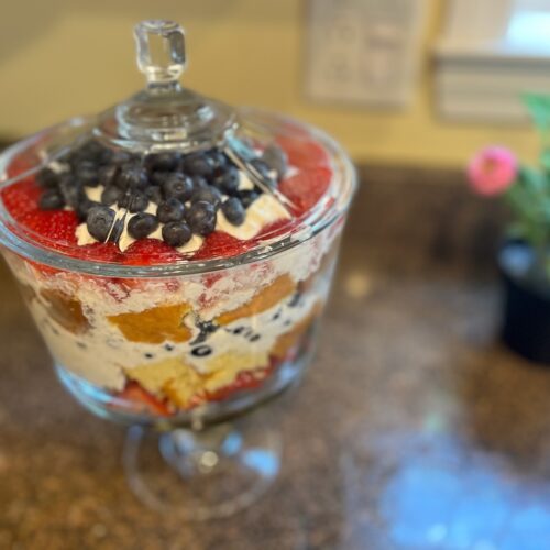 A glass trifle bowl filled with layers of cake, whipped cream, strawberries, and blueberries, placed on a granite countertop beside a potted pink flower.