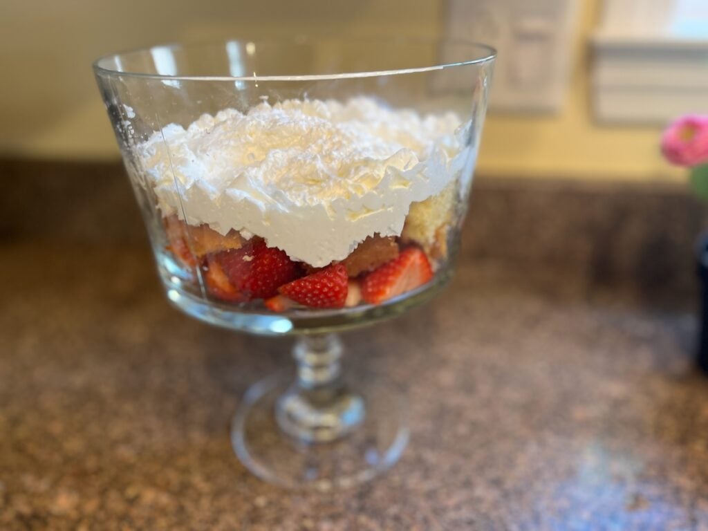 A glass trifle dish filled with layers of strawberries, sponge cake pieces, and whipped cream, sitting on a granite countertop.
