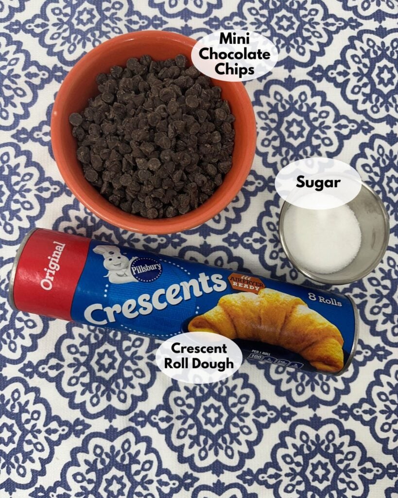 A bowl of mini chocolate chips, a small container of sugar, and a can of Pillsbury crescent roll dough on a blue and white patterned surface.