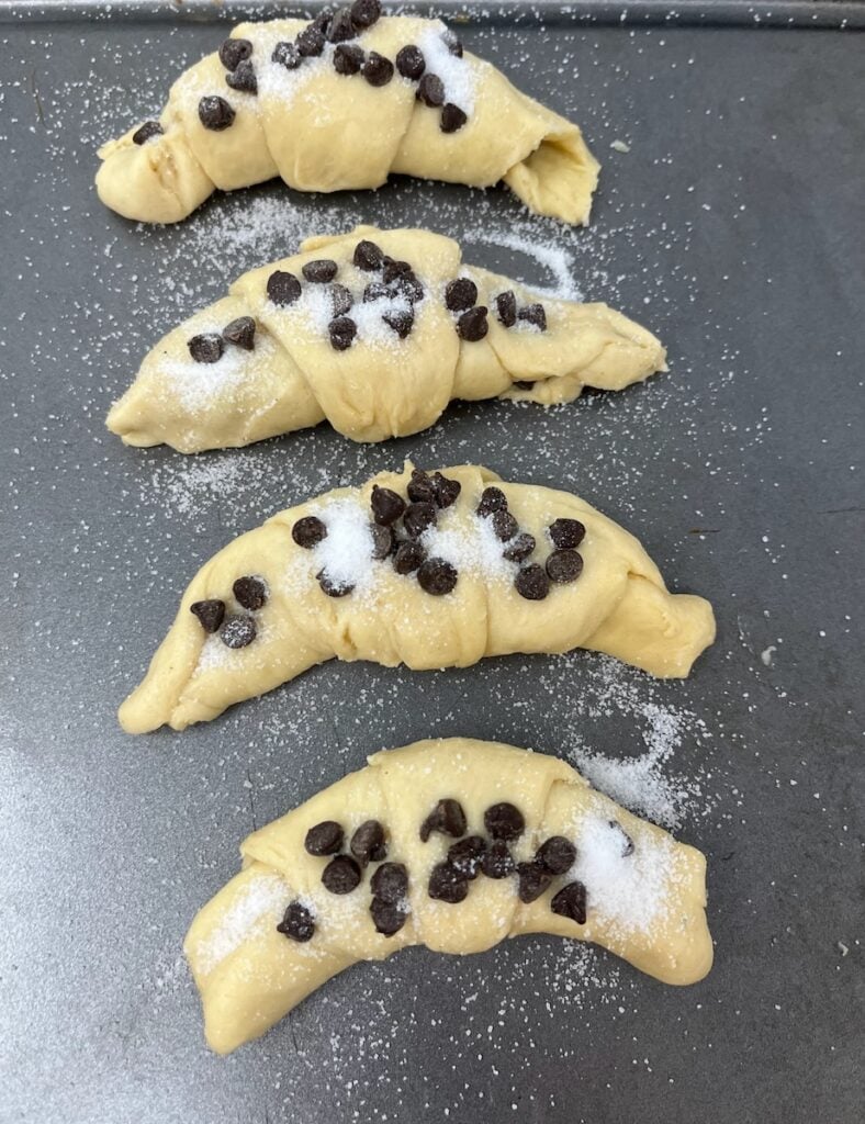Four uncooked crescent-shaped pastries topped with chocolate chips and sugar are placed on a dark baking tray.