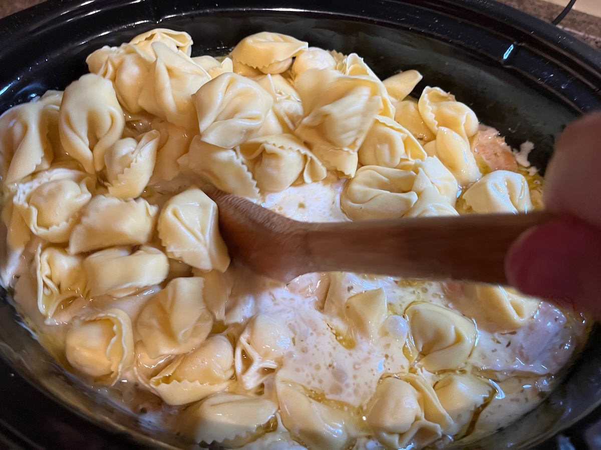 Close-up of tortellini pasta cooking in a creamy yellow sauce with pieces of chicken, stirred by a wooden spoon.