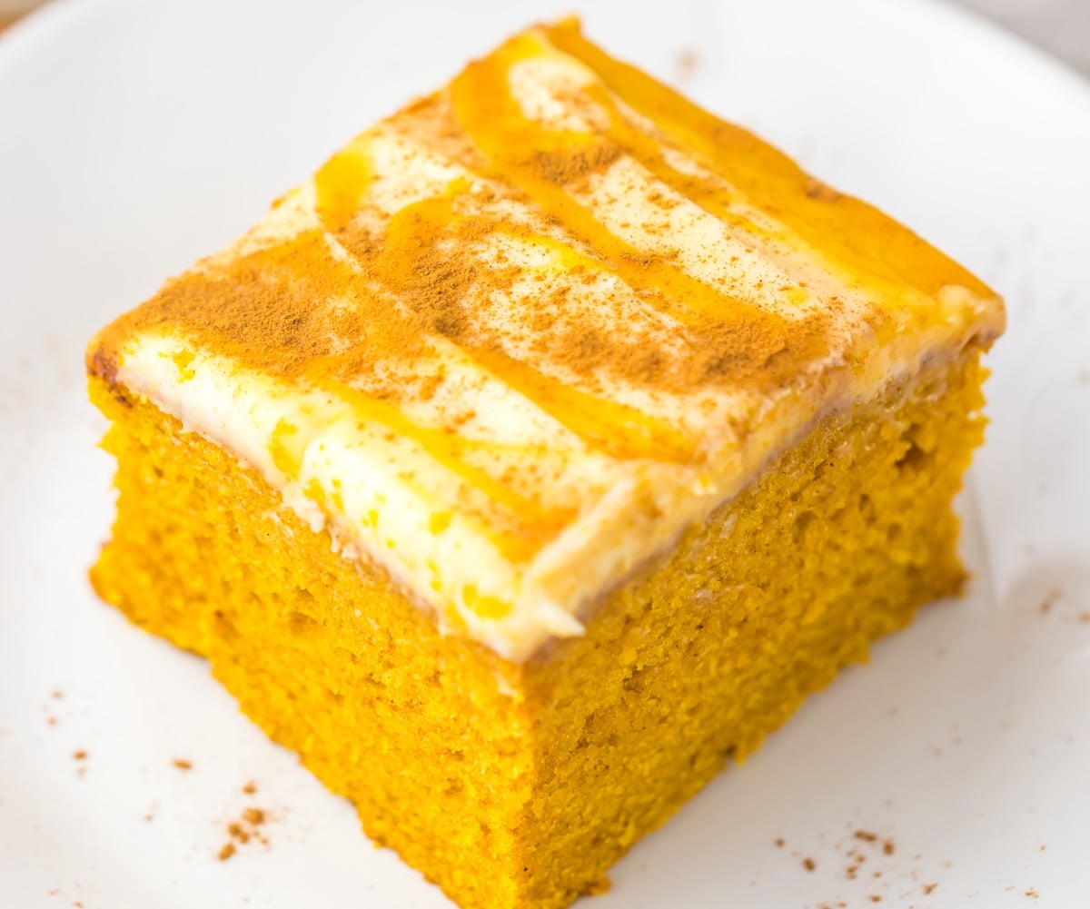 A square piece of orange-colored cake with a white frosting layer and orange drizzle on top, resting on a white plate.