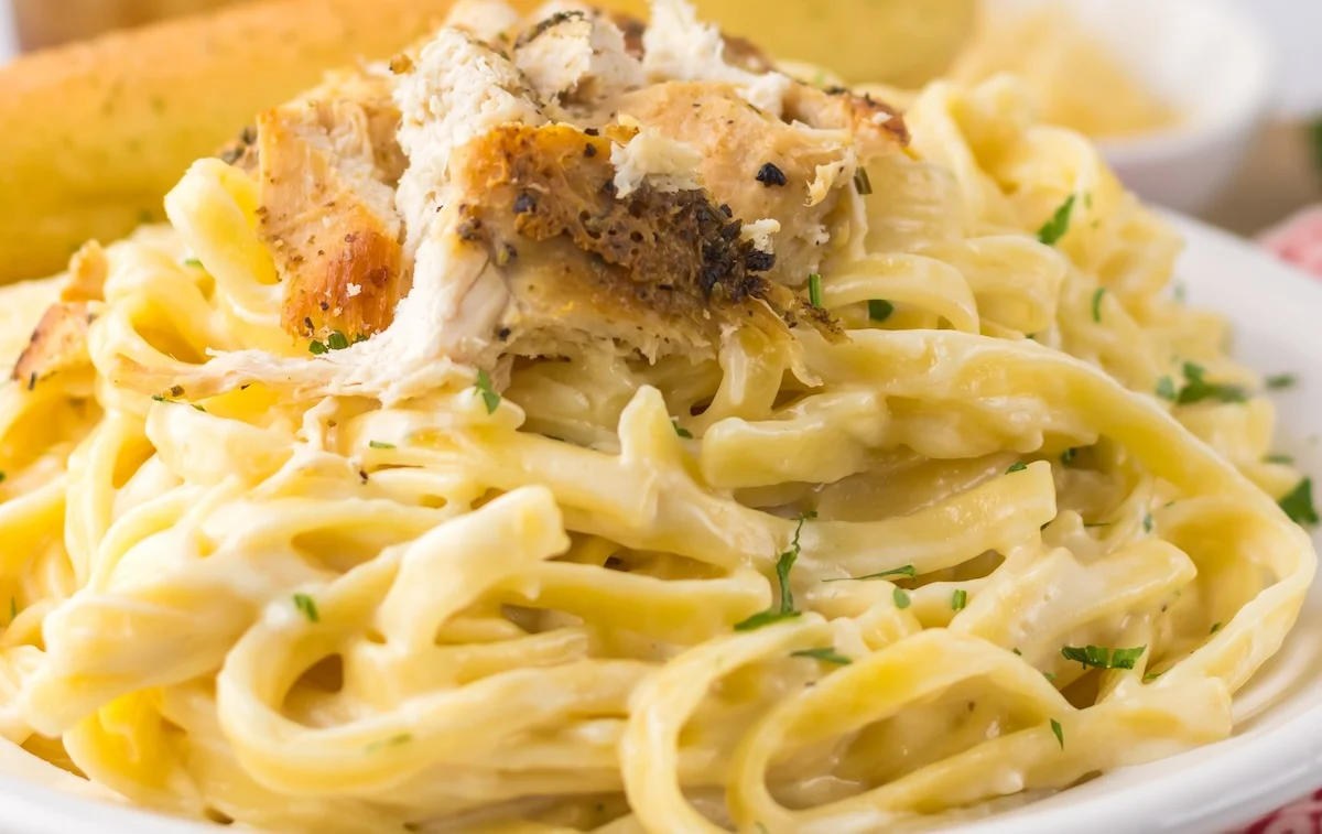 A plate of fettuccine Alfredo topped with pieces of grilled chicken and garnished with chopped herbs. A breadstick and a dipping sauce are visible in the background.