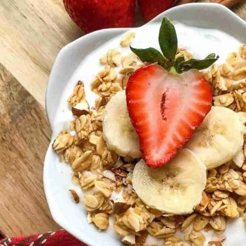 A bowl of yogurt topped with granola, banana slices, and a halved strawberry on a wooden board with a red cloth.
