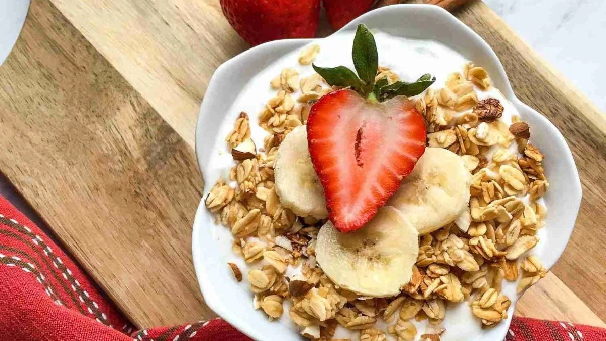 A bowl of yogurt topped with granola, banana slices, and a halved strawberry on a wooden board with a red cloth.