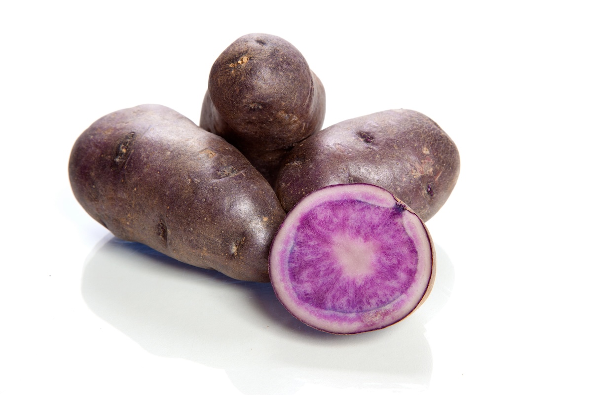 A group of three whole purple potatoes and one halved purple potato are displayed on a white surface. The interior of the halved potato is vibrant purple with a white ring.