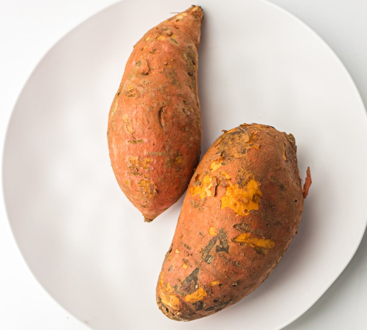 Two whole sweet potatoes with patches of dirt and rough skin placed on a white plate.