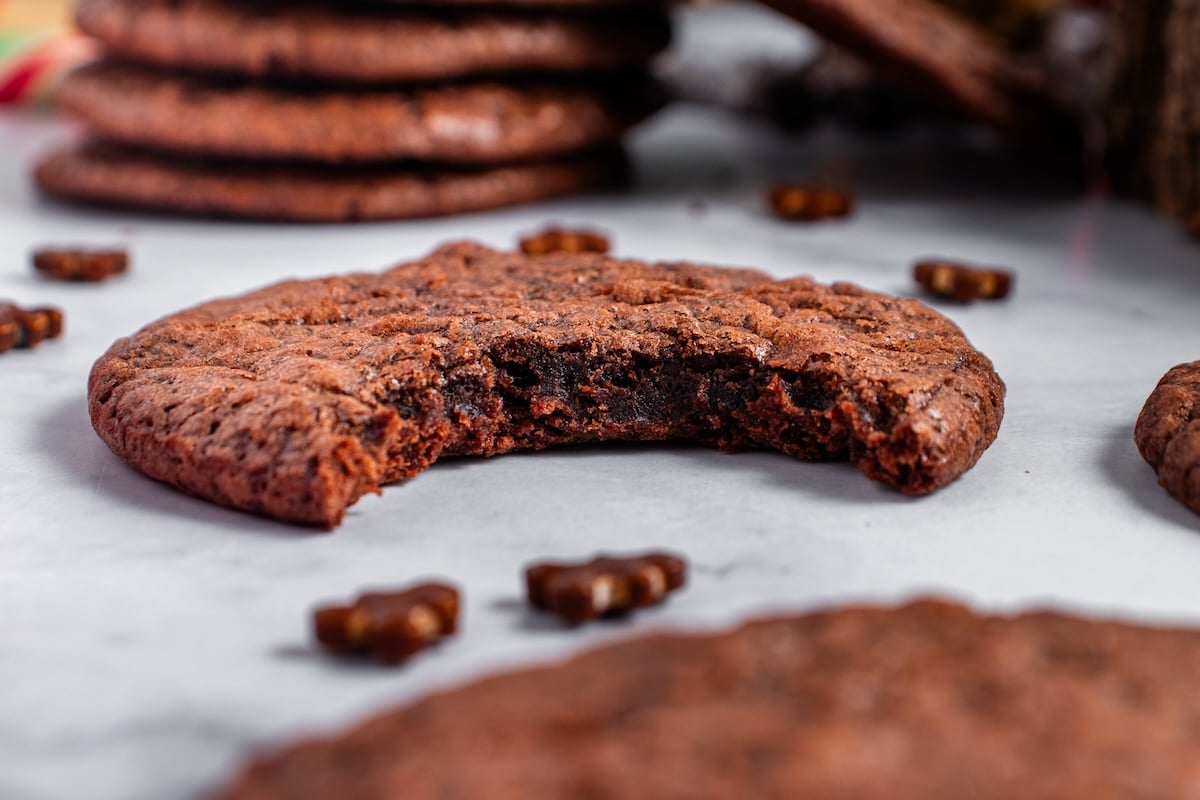 A close-up of a chocolate brownie cookie with a bite taken out reveals a gooey center. Small chocolate pieces are scattered around on the white surface, enhancing its irresistible allure.
