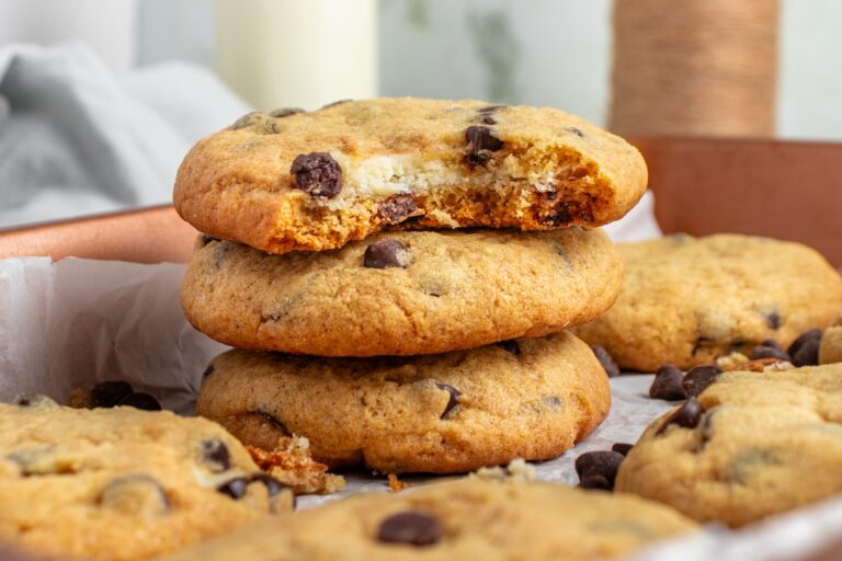 A stack of cheesecake stuffed cookies on parchment paper, with the top cookie missing a bite. Other cookies are scattered around.