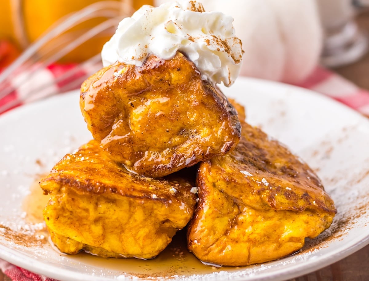 Three slices of Hawaiian Roll Pumpkin French toast topped with whipped cream and syrup on a white plate.
