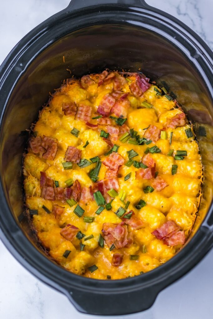 A slow cooker dish with melted cheese, bacon pieces, and chopped green onions on a bed of potatoes.