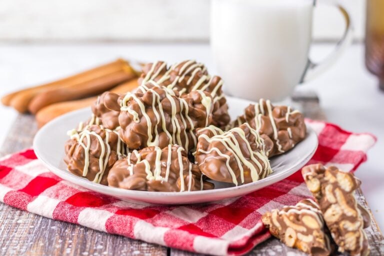 A plate of Crock Pot chocolate almond clusters drizzled with white chocolate sits on a red gingham napkin, perfectly paired with a glass of milk in the background.
