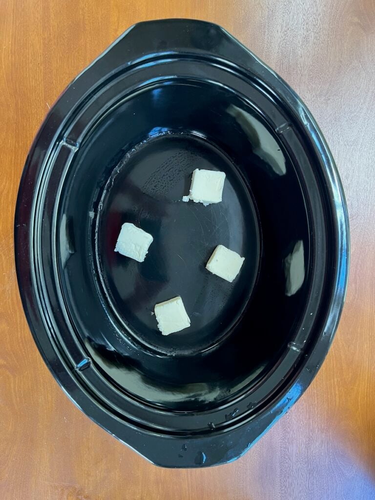 Four small white cubes are placed in a black Crock Pot, ready to transform into a delightful Hawaiian Roll French Toast Casserole on a wooden surface.