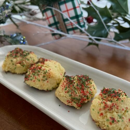Four festive cookies made from cake mix, adorned with red and green sprinkles, rest on a white platter with holiday decorations shimmering in the background.