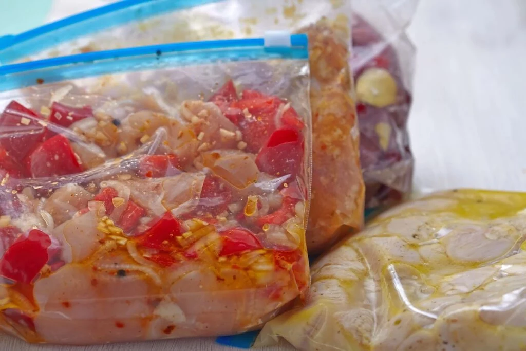 Three plastic bags with marinating ingredients, including diced vegetables and meats, are sealed and placed on a white surface, ready to become tasty freezer meals.