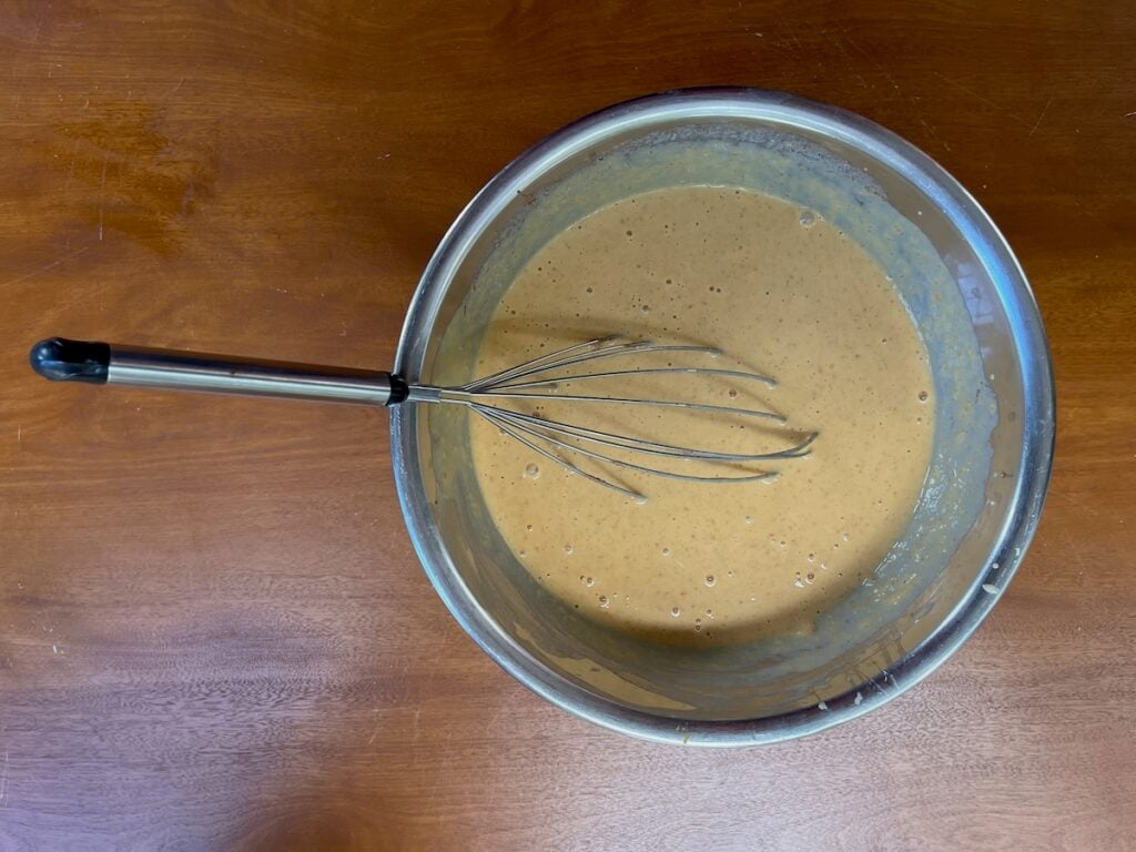 A metal bowl containing a whisk and tan-colored batter sits on a wooden surface, ready to become the perfect base for Crock Pot Hawaiian Roll French Toast Casserole.