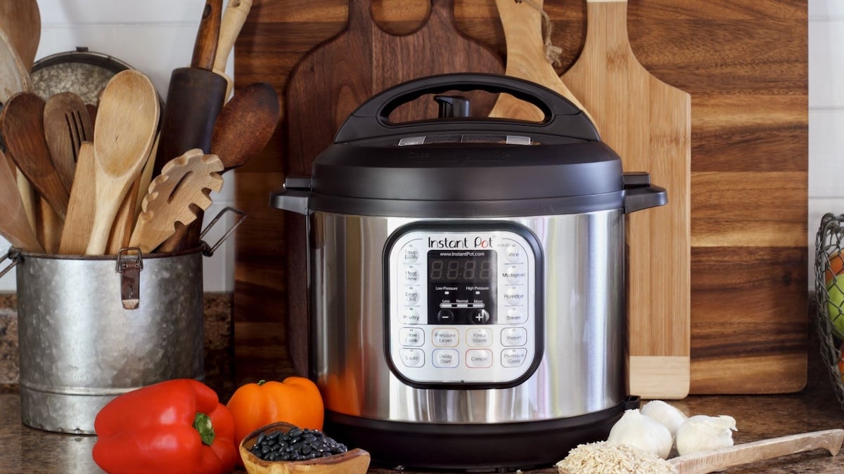 A silver multi-cooker sits on a kitchen counter surrounded by wooden utensils, cutting boards, a red bell pepper, an orange bell pepper, dry black beans, and uncooked rice.