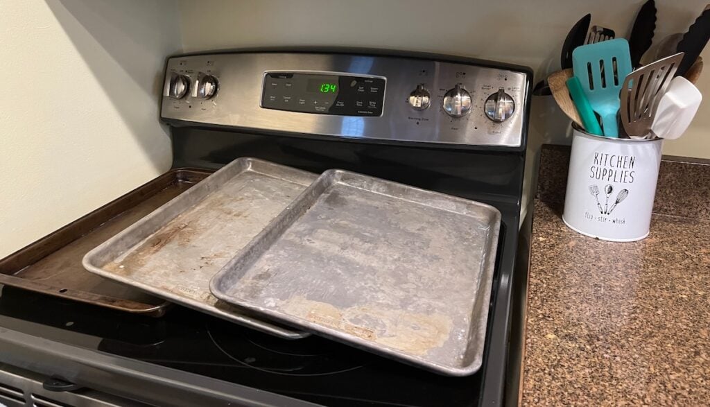 Two sheet pans rest on a stovetop next to a container labeled "Kitchen Supplies," brimming with utensils.