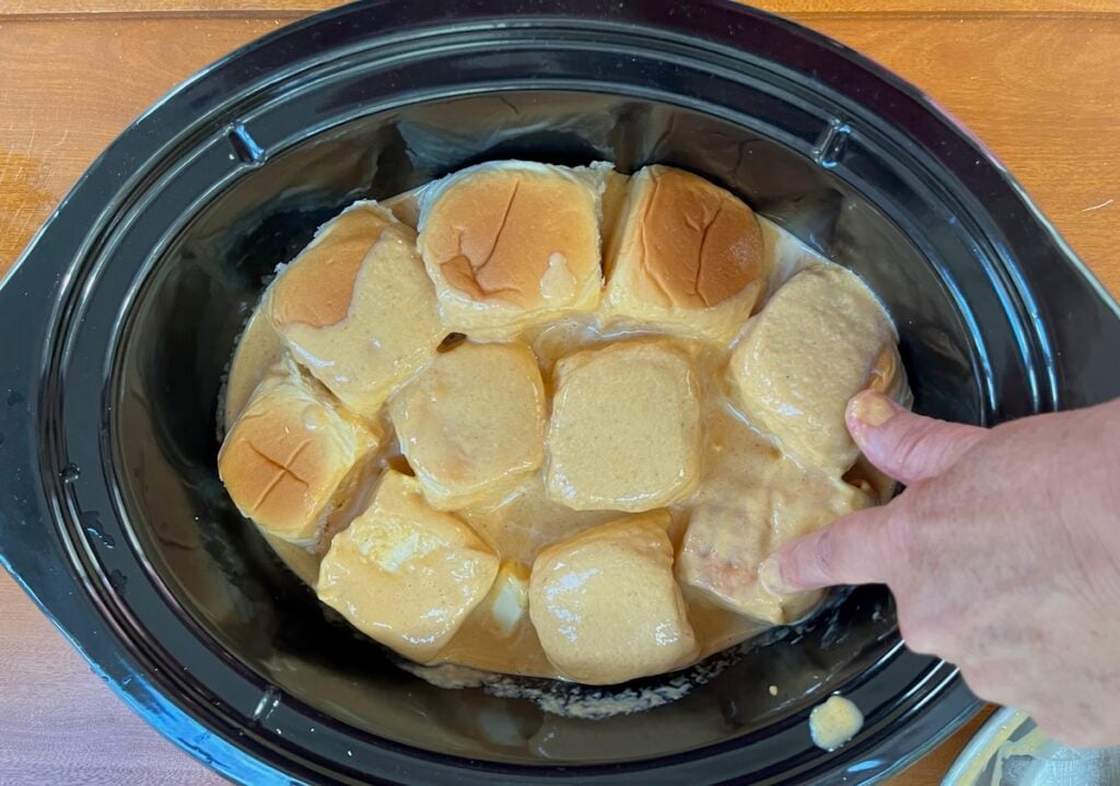A hand presses on Hawaiian rolls soaking in a creamy liquid inside a Crock Pot, creating a delightful Hawaiian Roll French Toast Casserole.