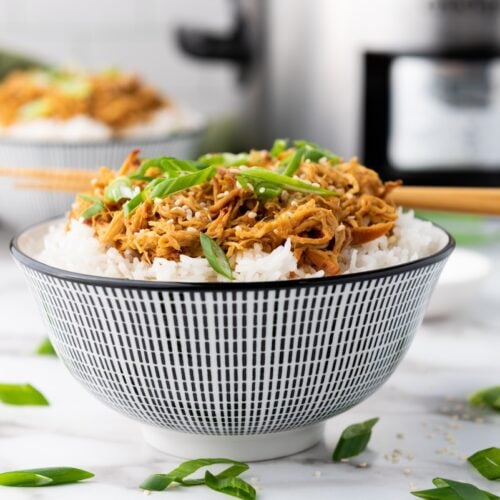 A bowl of white rice topped with Slow Cooker Honey Garlic Chicken and green onions, with chopsticks resting on top. Another similar bowl and a slow cooker are in the background.