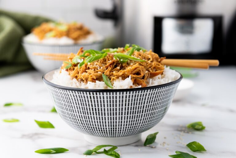 A bowl of white rice topped with Slow Cooker Honey Garlic Chicken and green onions, with chopsticks resting on top. Another similar bowl and a slow cooker are in the background.