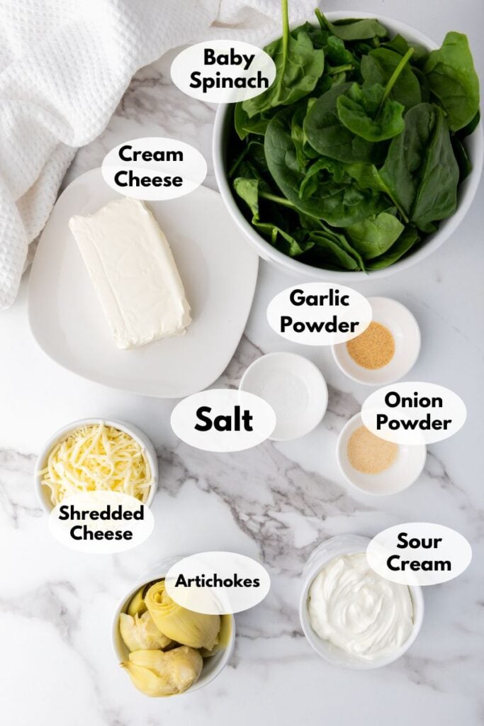 Top view of a cooking setup with bowls labeled: baby spinach, cream cheese, garlic powder, onion powder, salt, shredded cheese, artichokes, and sour cream on a marble surface.
