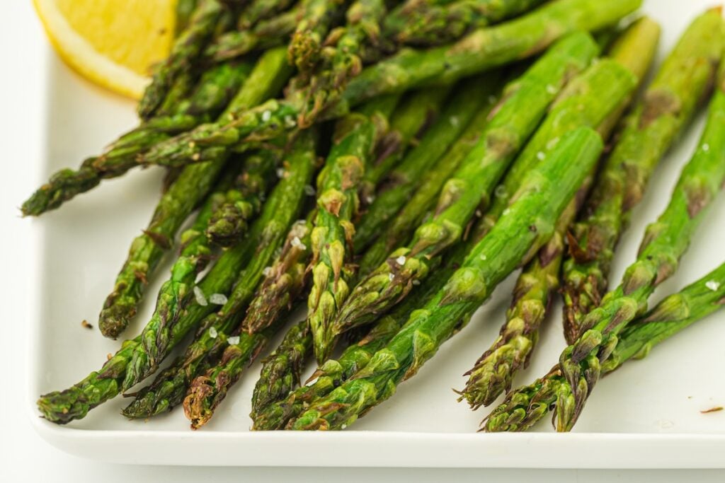 A plate of roasted asparagus spears garnished with sea salt, with a slice of lemon in the corner.