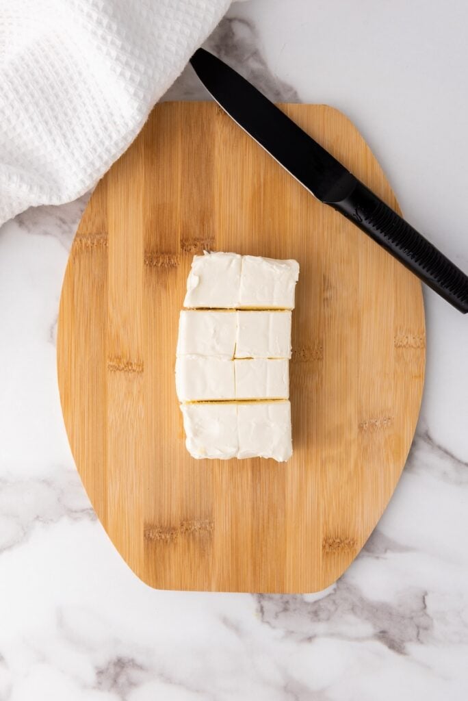 A block of cream cheese, destined for a delicious slow cooker spinach artichoke dip, is cut into cubes on a wooden cutting board with a black knife beside it.