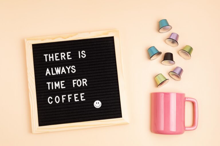 A letter board reads "There is always time for coffee" with a smiley face. A pink mug and scattered coffee capsules are nearby on a light beige background.