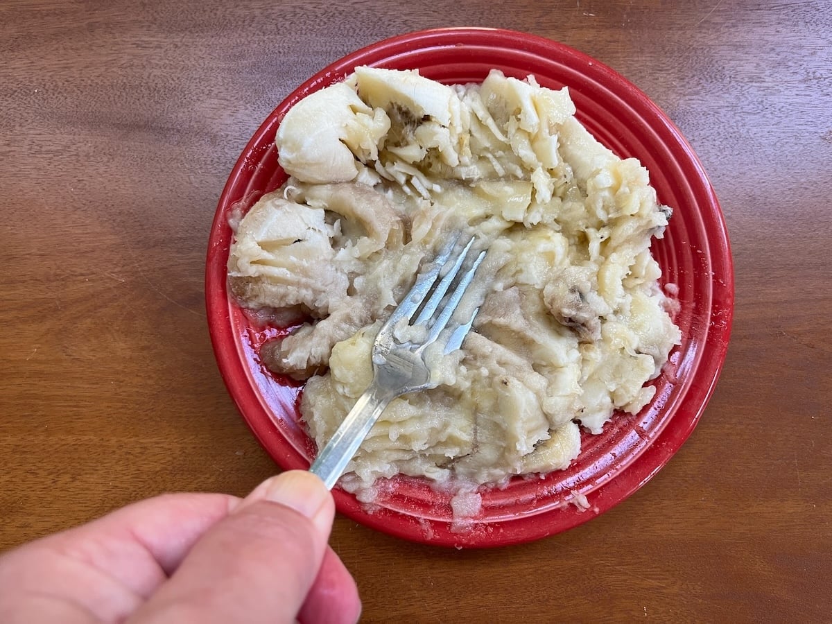 A hand holds a fork over a red plate of mashed bananas on a wooden table.