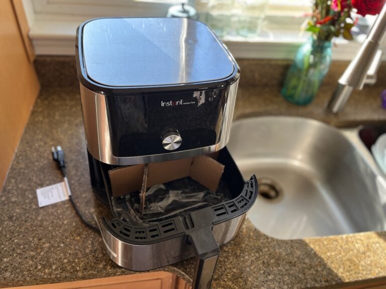 A modern air fryer sits proudly on the kitchen counter near a sink, its drawer open and packaging materials hinting at its first-time use.