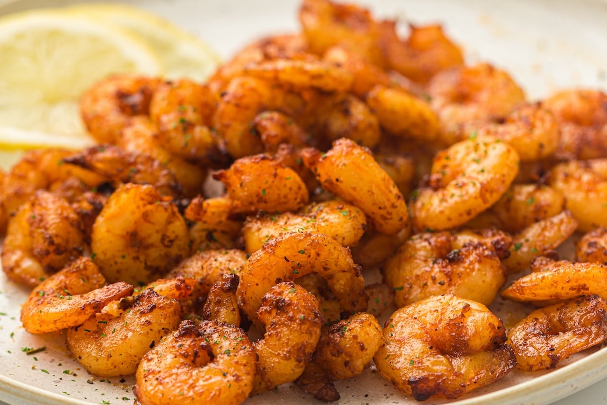 A plate of spicy, seasoned shrimp garnished with parsley, with lemon slices in the background.