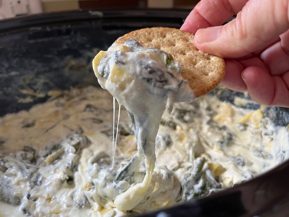 A hand dips a cracker into a rich, slow cooker spinach artichoke dip, unveiling layers of creamy goodness.