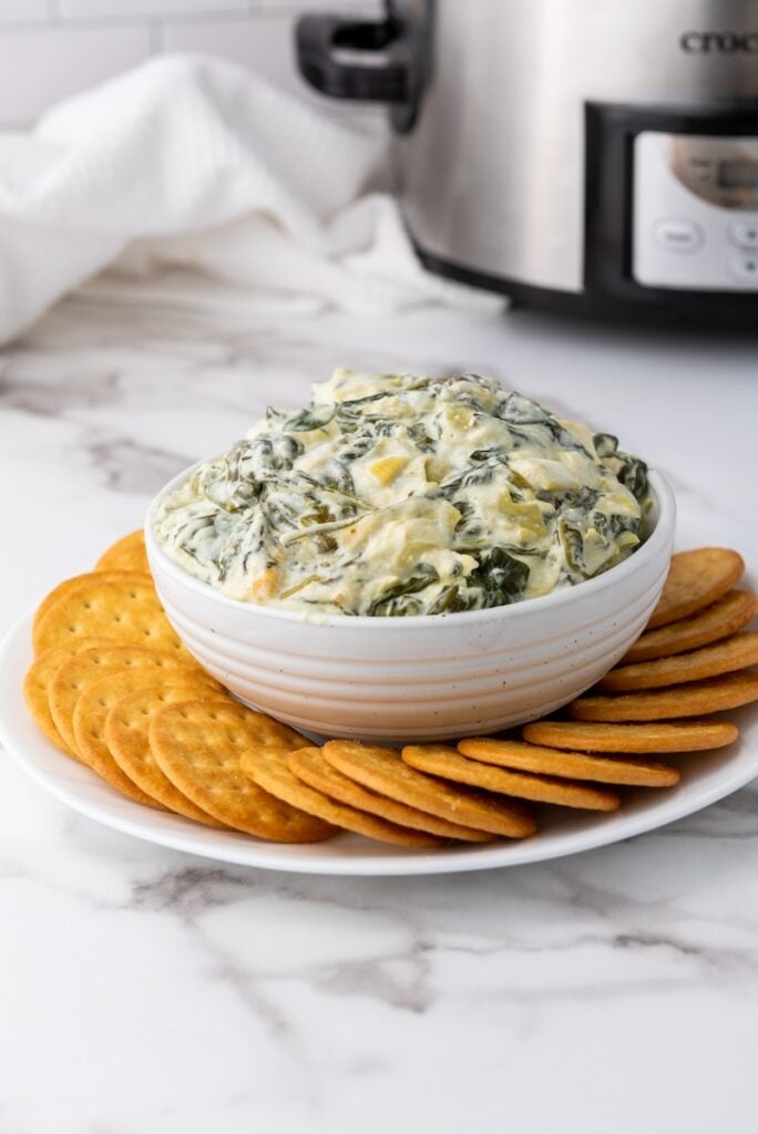 A bowl of creamy slow cooker spinach and artichoke dip sits on a plate surrounded by round crackers, with the trusty appliance gently steaming in the background.