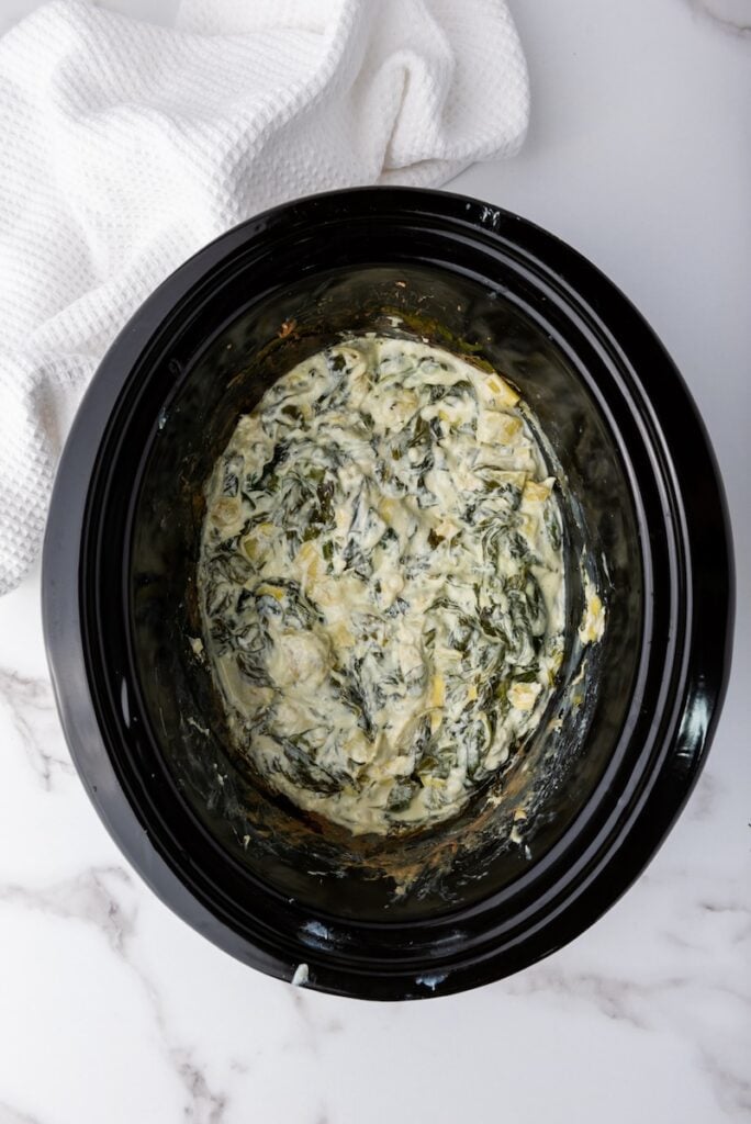 A black slow cooker containing creamy spinach artichoke dip sits on a marble surface, its rich aroma inviting. Nearby, a white cloth adds a touch of elegance to the scene.