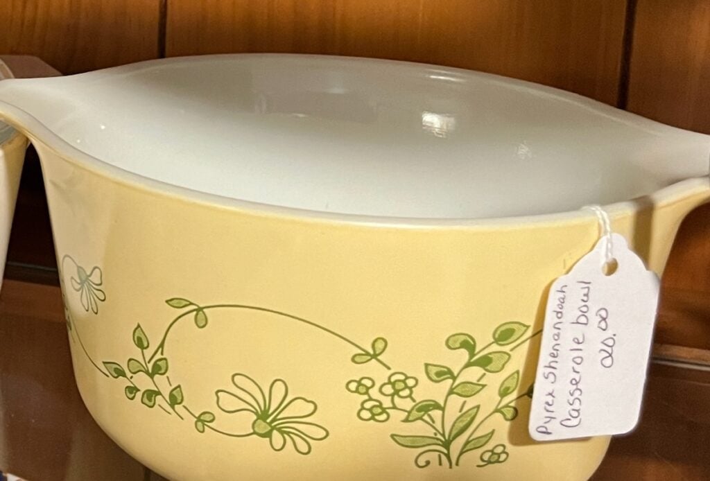 Vintage Pyrex Shenandoah casserole bowl with a green floral design, displayed on a shelf. A tag reading "Pyrex Shenandoah Casserole Bowl $6.00" is attached to the handle, accentuating its retro charm.