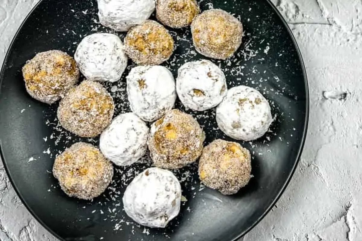 A black plate with round cookies coated in powdered sugar and shredded coconut on a textured gray surface.