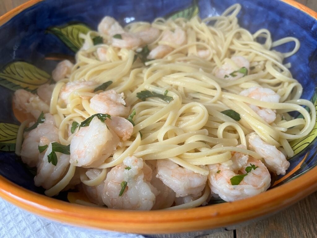 A bowl of linguine pasta with shrimp, garnished with parsley, in a blue and orange bowl on a wooden table.