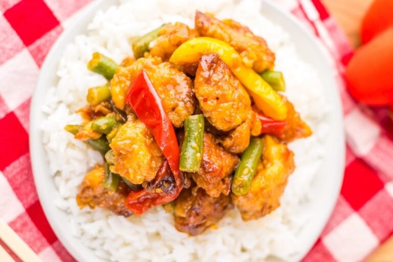 A plate of white rice topped with stir-fried chicken, bell peppers, and green beans, set on a red checkered tablecloth.